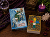 Thoth Tarot Premier Edition box lying flat beside a stacked deck showing the multicolored geometric card back, surrounded by a purple candle, crystal sphere, quartz cluster, greenery, and ritual décor.