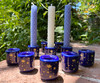 Several starry night cobalt and gold mini candle holders on brick with greenery in the background, arranged in a triangle, with three in the background holding beeswax handmade mini candles. 