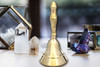 One Wiccan brass altar bell stands on a white table with crystals and a votive candle holder in the background. Bells are used for sound cleansing, transmuting energy, ritual, and goddess energy.