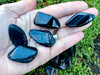 Several blue tiger eye tumbled stones lie on an open palm with a bed of greenery in the background displaying variations in color, size, and shape.