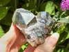 Glittering Peruvian pyrite cluster with matrix held between fingers, glowing in the sunlight with greenery behind. Revered for its ability to attract wealth and promote strong, protective energy.