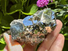 Peruvian pyrite cluster held between fingers in direct sunlight, showing off a vivid golden sparkle against a backdrop of greenery. A powerful stone for boosting self-worth and shielding energy fields.
