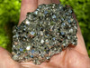 A single high-quality Peruvian pyrite cluster held in hand with greenery behind, sparkling brilliantly in filtered sunlight. This stone enhances willpower, confidence, and energetic protection.