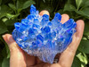 Close-up of large Tanzine Aura Quartz cluster held between fingers in sunlight with greenery, blue violet aura quartz for chakra alignment.