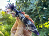 Chevron Dream Amethyst Athame with star-crowned handle held between fingers, multiple crystal athames in background on greenery, natural chevron amethyst patterns visible.