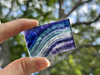 Polished rainbow fluorite slab held between two fingers, background of greenery highlighting wavy bands of purple, blue, white, and teal fluorite. Polished rainbow fluorite slab held between two fingers, background of greenery highlighting wavy bands of purple, blue, white, and teal fluorite.