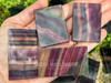 Open palm holding four rainbow fluorite slabs in sunlight, capturing variations in color and banding with purple, clear, green, blue, and yellow crystal hues. Open palm holding four rainbow fluorite slabs in sunlight, capturing variations in color and banding with purple, clear, green, blue, and yellow crystal hues.
