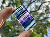 Polished rainbow fluorite slab held between fingers in sunlight with greenery background, highlighting vibrant stripes of clear, purple, and blue. Polished rainbow fluorite slab held between fingers in sunlight with greenery background, highlighting vibrant stripes of clear, purple, and blue.