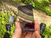 Rainbow fluorite polished slab resting between cupped fingers in sunlight, with more slabs on greenery, showing curved lines in purple, green, and clear tones. Rainbow fluorite polished slab resting between cupped fingers in sunlight, with more slabs on greenery, showing curved lines in purple, green, and clear tones.