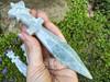 Moss agate crystal athame held in a cupped hand with more in the background on greenery and rock, displaying smooth polish. Moss agate crystal athame held in a cupped hand with more in the background on greenery and rock, displaying smooth polish.