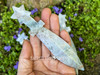 Moss agate crystal athame resting on an open palm with more in the background on greenery and rock, showing color and texture. Moss agate crystal athame resting on an open palm with more in the background on greenery and rock, showing color and texture.