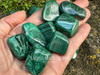 Assorted malachite tumbled stones resting across an open palm with rock and greenery background, crystals for heart chakra healing.