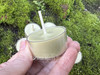 Side view of a real bayberry tealight held between fingers, displaying depth and the clean-burning bayberry and beeswax blend with more tealights resting on moss-covered brick behind it.