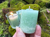 One Sweetgrass & Cedar Cleansing Votive Candle rests between fingers to show size with more on moss covered bricks behind it and one candle lit in a clear glass votive cup.
