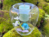 One floating handblown glass votive and tealight candle holder rests on moss-covered brick. Inside the candle cup rests a lit sweetgrass and himalyan cedar votive candle below it charging are crystals and flowers.