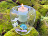 One floating handblown glass votive and tealight candle holder rests on moss-covered brick. Inside the candle cup rests our sweetgrass and himalyan cedar votive candle below it charging are crystals and flowers.