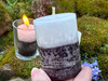 One Reversing Black and White Votive Candle rests between fingers to show size with more on moss covered bricks behind it and one candle lit in a clear glass votive cup.

