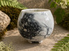 Black and white handmade glazed ceramic incense bowl with three-footed base displayed outdoors among moss and natural stone in bright sunlight. Shown from the front. 
