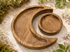Empty wooden crescent moon and round altar tray set displayed outdoors on natural stone surrounded by greenery and wildflowers.