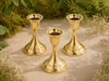 Three polished brass taper candle holders for standard taper candles arranged outdoors on natural stone with greenery and white flowers in bright natural sunlight.