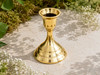 Polished brass taper candle holder with stepped base for standard taper candles displayed outdoors on natural stone surrounded by greenery and delicate white flowers in warm sunlight.