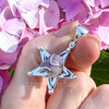Angled close-up of a natural Grape Chalcedony pentagram pendant held against pink and purple flowers, highlighting the crystal texture and silver star design.