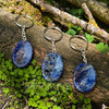 Three natural Lapis Lazuli worry stone keychains with silver-colored hardware displayed on a mossy log, showing polished blue stones with naturally varied white and dark mineral patterns.