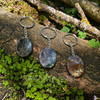 Three natural Moss Agate worry stone keychains with silver-colored hardware displayed on a mossy woodland log, showing varied green, gray, brown, and translucent patterns.