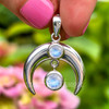 Rainbow Moonstone sterling silver crescent pendant held upright, showing the centered cabochon and freely hanging Moonstone with blue flash.