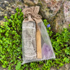 White sage, palo santo, and amethyst smudge bundle set packaged in a sheer drawstring bag and displayed against mossy stone and greenery.
