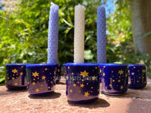 A close up front view of several starry night cobalt and gold mini candle holders on brick with greenery in the background, arranged in a triangle, with three in the background holding beeswax handmade mini candles. 