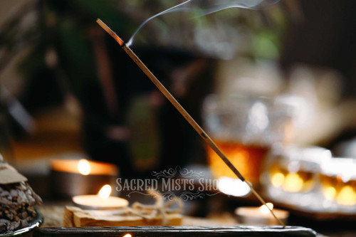 Triloka  Frankincense, Myrrh, Sandalwood Premium Masala Stick Incense is burning on a black ash catcher with candles in the background