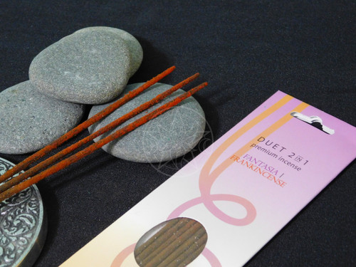 A close-up of one package of Frankincense Fantasia Duet Incense by Triloka lying on a black altar cloth with several smooth rocks and a round silver mandala inense holder showing three sticks of incense lying next to the package. Handmade incense with a fruity bouquet followed by the balsamic scent of frankincense ideal for daily use and meditation. 10 sticks per pack.