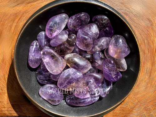 a close up of a bronze bowl filled with ametrine in the sunlight displays variations in coloring, shape, markings, and size. Ametrine tumbled stone blends the uplifting clarity of citrine with the spiritual insight of amethyst, making it perfect for enhancing focus, creativity, and inspired action.