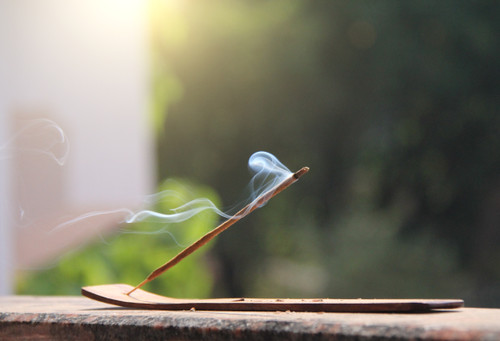 Nag champa incense stick burning on a wood ash catcher on a brick ledge with greenery in the background. Nag Champa Premium Masala Incense Sticks by Satya Sai Baba 40 grams for Offerings, Spells, Rituals, Meditation, Aromatherapy, Calming.