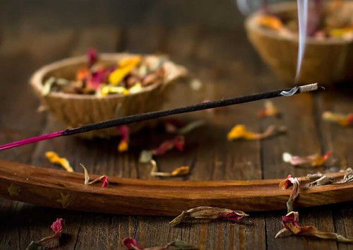 precious lavender incense stick burning on a wooden ash catcher with rose petals strewn over a wood table and bowls.