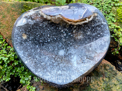 one druzy agate crystal heart standing on mossy bricks with greenery in the background shown from the bottom angled