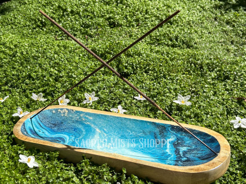Double Stick Incense Burner Trough Style with an ocean design is lying on a bed of greenery with white flowers, showing two incense sticks in the burner.