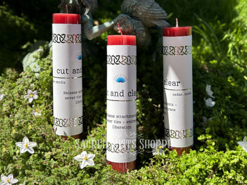 Three cord cutting red pillar spell candles scented with essential oils of cedar and clove are standing in greenery and white flowers with wood in the background. Perfect for cutting cords, releasing attachments, letting go, and severing ties.