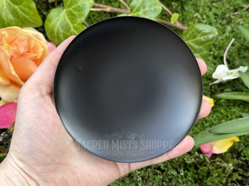 One black iron candle plate is held in hand to display size, and a bed of greenery with orange, pink, and white flowers is behind it.