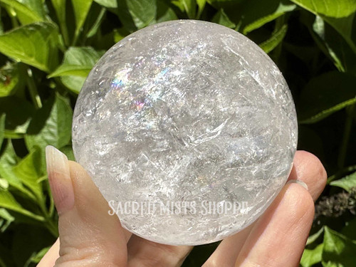 A hand is holding a premium 50mm+ Lemurian Quartz crystal sphere in the sunlight with lush greenery in the background; this high-vibration stone enhances spiritual connection, inner wisdom, and intuitive insight. Each sphere includes a free wooden stand, making it perfect for energy work, meditation, or display.