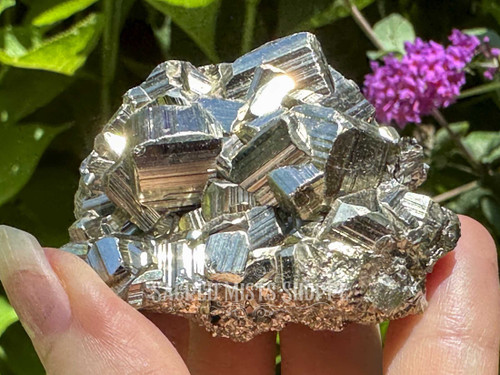 Ethically sourced cubic Peruvian pyrite cluster held between fingers, sparkling in the sunlight with lush greenery in the background. Known for attracting abundance and shielding negative energy.