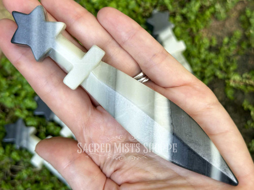 Zebra agate crystal athame with star-crowned handle resting across an open palm, with more athames in the background on greenery.