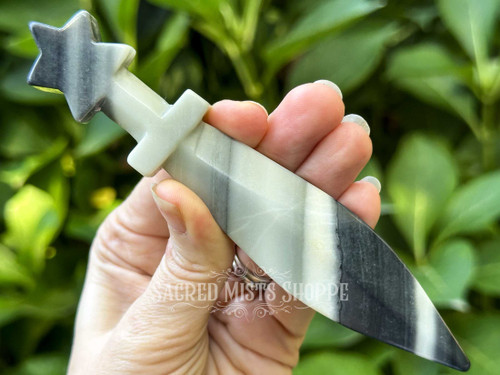 Zebra agate athame held between fingers in a cupped hand, greenery in the background highlighting black and white banding. Zebra agate athame held between fingers in a cupped hand, greenery in the background highlighting black and white banding.