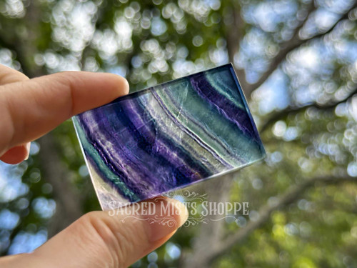 Rainbow fluorite crystal slab held between fingers in sunlight with greenery behind, showing clarity and flowing bands of purple, clear, blue, and yellow hues. Rainbow fluorite crystal slab held between fingers in sunlight with greenery behind, showing clarity and flowing bands of purple, clear, blue, and yellow hues.