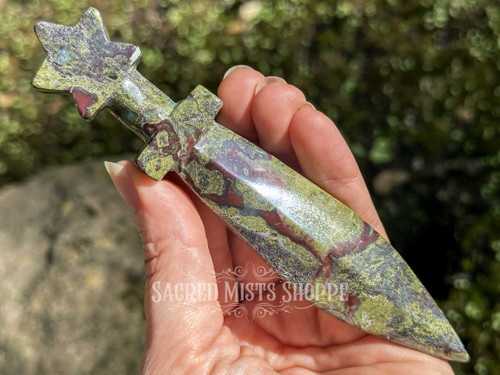 Dragon’s blood jasper athame held between fingers on a cupped palm with greenery and rock in the background, highlighting stone detail and craftsmanship.