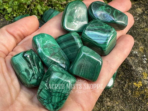 Assorted malachite tumbled stones resting across an open palm with rock and greenery background, crystals for heart chakra healing. Assorted malachite tumbled stones resting across an open palm with rock and greenery background, crystals for heart chakra healing.