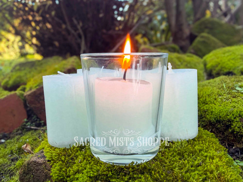 several sage bundle handmade votive candles sit on moss covered brick, with one in the foreground in a clear votive cup while lit displaying the candle and its beauty.