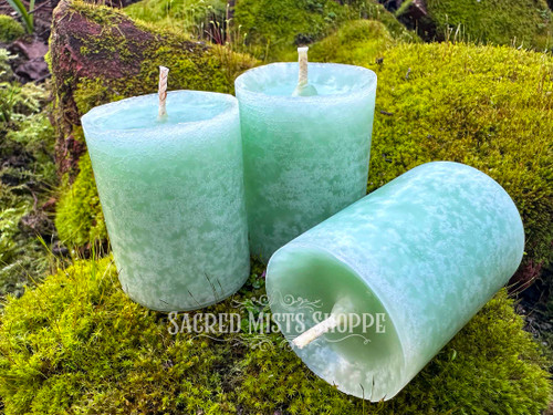 Three Sweetgrass & Cedar Cleansing Votive Candles are shown from the front and stand on moss covered bricks and one rests on the side.