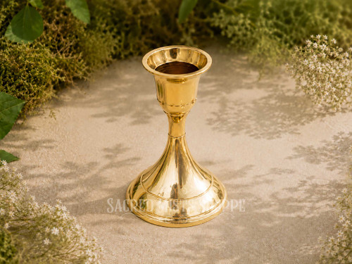 Polished brass taper candle holder for standard taper candles displayed outdoors on natural stone surrounded by greenery and delicate white flowers in warm sunlight.